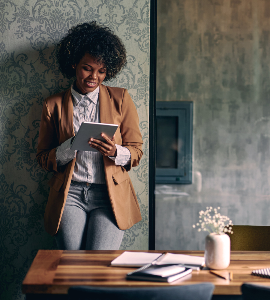 Decision Excellence Homepage Image Smiling woman in a blazer leaning against a wall, using a tablet in a warmly lit room with a wooden table and notebooks in the foreground.