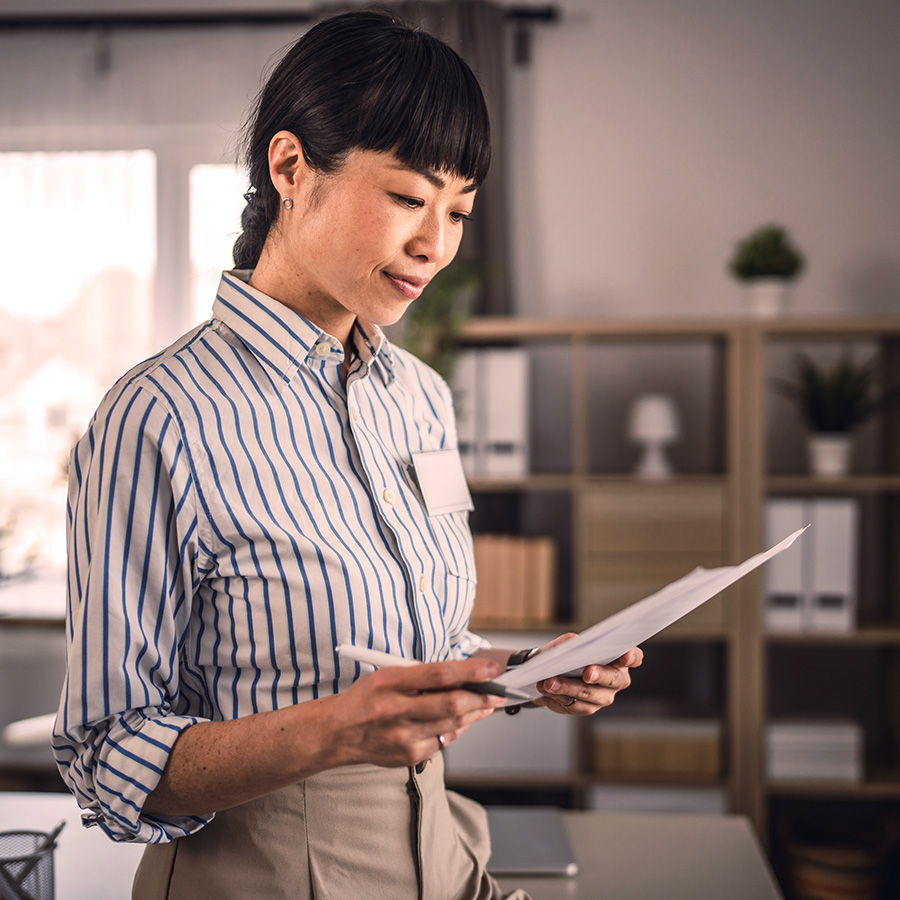 Decision Excellence Japanese Homepage Image Professional woman in a striped shirt reviewing documents in a modern office setting, surrounded by shelves and natural light.