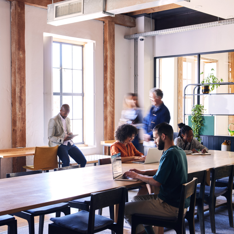 Homepage Hero Image Modern coworking space with people working on laptops and tablets at a large wooden table, surrounded by natural light and greenery.
